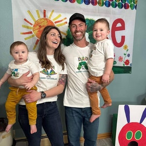 Puede incluir: Una familia de cuatro personas con camisetas blancas con el texto "Soy la oruga muy hambrienta" y una imagen de la oruga. El padre lleva a un ni&ntilde;o en brazos. La madre lleva a un ni&ntilde;o en brazos. La familia est&aacute; de pie frente a un tel&oacute;n de fondo colorido con un sol y la letra "e".