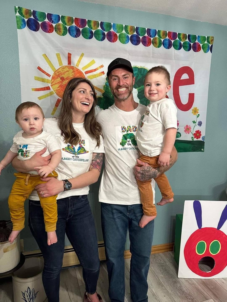 Puede incluir: Una familia de cuatro personas con camisetas blancas con el texto "Soy la oruga muy hambrienta" y una imagen de la oruga. El padre lleva a un ni&ntilde;o en brazos. La madre lleva a un ni&ntilde;o en brazos. La familia est&aacute; de pie frente a un tel&oacute;n de fondo colorido con un sol y la letra "e".