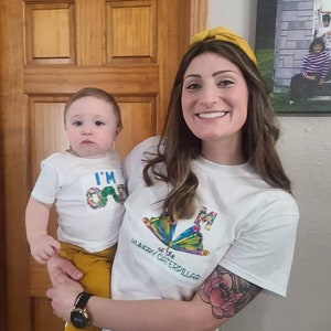 Puede incluir: Una mujer y un ni&ntilde;o con camisetas blancas a juego con una mariposa y el texto "I'm One" y "I'm the Hungry Caterpillar". La mujer lleva una diadema amarilla y tiene un tatuaje en el brazo.