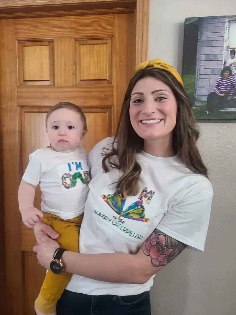 Puede incluir: Una mujer y un ni&ntilde;o con camisetas blancas a juego con una mariposa y el texto "I'm One" y "I'm the Hungry Caterpillar". La mujer lleva una diadema amarilla y tiene un tatuaje en el brazo.