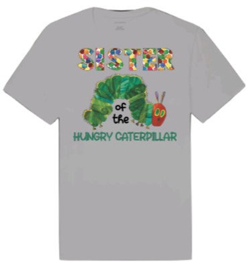 Puede incluir: Camiseta gris con un gr&aacute;fico colorido de "Hermana de la Oruga Hambrienta". La oruga es verde con una cabeza roja y est&aacute; rodeada de flores coloridas.