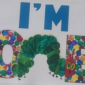 Puede incluir: Una pancarta de cumplea&ntilde;os colorida con las palabras "I'M ONE" en letras azules. Las letras O y E est&aacute;n hechas de c&iacute;rculos coloridos, y la letra N es una oruga verde.