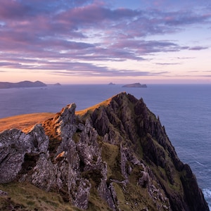 Puede incluir: Un acantilado rocoso que domina un vasto océano con un cielo morado y rosa. El acantilado está cubierto de hierba verde y rocas marrones. En la distancia, se pueden ver varias islas pequeñas.