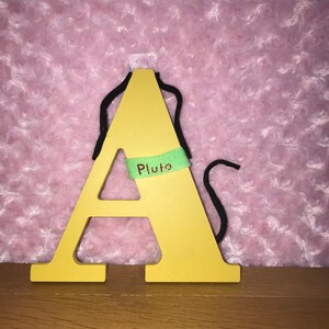 May include: A yellow wooden letter A with a green tag that says "Pluto" hanging from a black string.