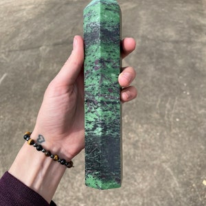May include: A green and black gemstone point, also known as a crystal, with a faceted tip. The point is about 6 inches long and is held in a person's hand.