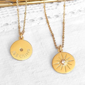Pode incluir: Dois colares de ouro com pingentes. Um pingente é um disco redondo com o texto "J & B" e um símbolo de coração. O outro pingente é um sol com um pequeno diamante no centro.