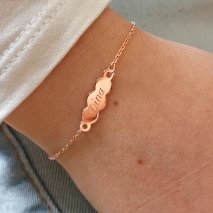 May include: A rose gold bracelet featuring a chain and a central, elongated charm. The charm is engraved with the name "Dina". The bracelet is displayed on a wrist, indicating it's a piece of jewellery.