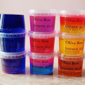 Puede incluir: Seis pequeños recipientes de plástico transparente llenos de gel de ducha de colores. Cada recipiente tiene una etiqueta negra con el texto "Olive Roe" y una descripción del aroma del gel de ducha. Los aromas son Bayas Inglesas, Sorbete de Fresa, Algodón de Azúcar, Limonada Rosa, Tutti Frutti y Mango & Papaya.