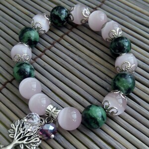 May include: A bracelet made of pink and green beads with a silver tree charm. The beads are alternating colors and have silver accents. The tree charm has a small purple gem.
