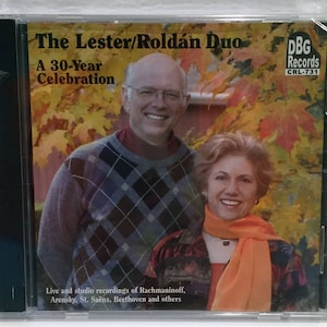 May include: A CD cover featuring a couple, a man and a woman, smiling at the camera. The background is a blurred image of autumn leaves. The title of the CD is "The Lester/Roldán Duo: A 30-Year Celebration". The CD is from DBG Records, CRL-731. The text on the CD cover reads "Live and studio recordings of Rachmaninoff, Arensky, St. Saëns, Beethoven and others."