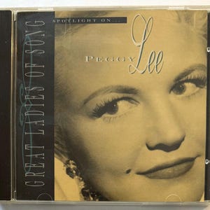 Puede incluir: Una caja de CD con una imagen en tonos sepia de Peggy Lee. El texto dice "Great Ladies of Song" y "Spotlight On... Peggy Lee". La portada muestra un primer plano del rostro de Lee, con su nombre en una elegante escritura.