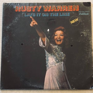 May include: A vinyl record album cover featuring a woman in a sequined dress, pointing with her right hand. The album title is "Rusty Warren Lays It On The Line" and the record label is "Cresendo".