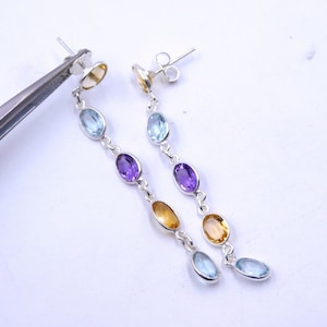 Puede incluir: Un par de pendientes de plata con un diseño graduado que presenta piedras preciosas ovaladas en tonos azules, morados y amarillos.