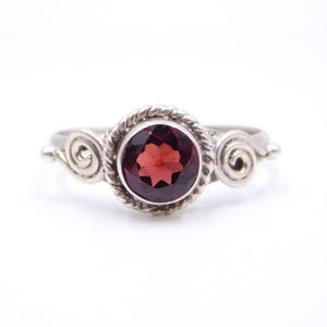 Può includere: Anello d'argento con una gemma rosso scuro al centro, circondata da un motivo a corda intrecciata. L'anello presenta dettagli decorativi a spirale su entrambi i lati della gemma. Un gioiello.
