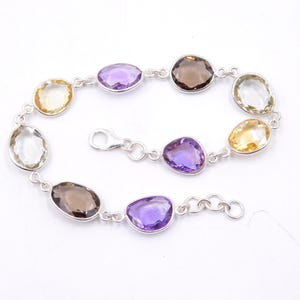 Peut inclure: Un bracelet en argent orné de pierres précieuses ovales de différentes couleurs : violet, jaune, marron et transparent. Les pierres sont reliées par des maillons en argent et un fermoir.