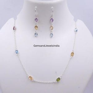 Puede incluir: Un conjunto de collar y pendientes de plata con piedras preciosas ovales en varios colores, incluyendo púrpura, amarillo y azul. El conjunto se exhibe sobre un busto blanco. El texto "GemsandJewelsIndia" es visible.