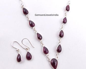 May include: A silver necklace and earring set featuring teardrop-shaped, deep red gemstones. The necklace has a pendant design, and the earrings are simple drops. The text "GemsandJewelsIndia" is visible.