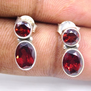 May include: A pair of silver earrings with red gemstone drops. The earrings feature two round gemstones and one oval gemstone.