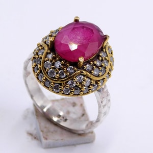 以下が含まれることがあります： 大きな楕円形のルビーを中央に配したシルバーリング。ルビーは深い赤色で、小さな透明な宝石で囲まれています。リングは、金色のアクセントが施された、詳細で装飾的なデザインです。ジュエリーです。