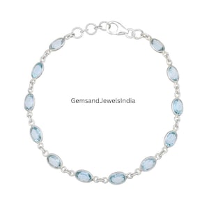 May include: A silver bracelet with oval, pale blue gemstones. The gemstones are set along the chain. The bracelet has a clasp fastening. The text "GemsandJewelsIndia" is visible.
