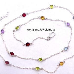 May include: A silver necklace with a fine chain, set with colourful gemstones. The necklace displays a variety of round-cut gems in red, green, yellow, blue, and purple. The clasp is visible. The text "GemsandJewelsIndia" is also visible.