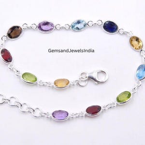 May include: A silver bracelet with oval gemstones in shades of red, purple, yellow, and blue. The gemstones are connected by a delicate chain and a secure clasp. The bracelet is presented on a white background.