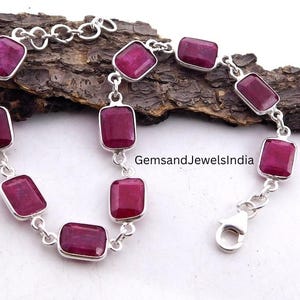 Può includere: Un bracciale in argento con pietre preziose rettangolari rosso scuro. Le pietre sono incastonate in singole montature d'argento e collegate da maglie d'argento. Il bracciale è esposto su un pezzo di legno scuro. Il testo "GemsandJewelsIndia" è visibile.