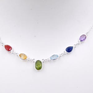 Op de afbeelding: Een zilveren ketting met een reeks gekleurde edelstenen in druppelvorm. De edelstenen zijn gerangschikt in een regenboogspectrum, waaronder rood, oranje, geel, groen, blauw en paars. De ketting wordt gepresenteerd tegen een witte achtergrond.