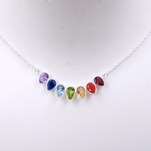 Puede incluir: Un collar de plata con una serie de piedras preciosas en forma de lágrima en colores del arcoíris. Las piedras están dispuestas en una línea curva, desde el morado hasta el rojo. Se muestra sobre un fondo blanco.