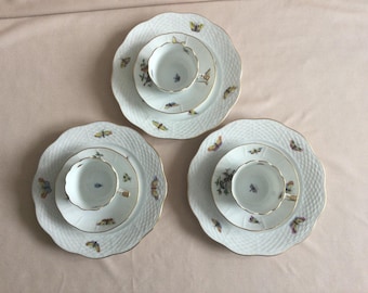 May include: Three white porcelain tea sets with butterfly and floral designs. Each set includes a teacup, saucer, and a larger plate. The plates have a textured, woven pattern and gold trim. The teacups and saucers are decorated with butterflies.