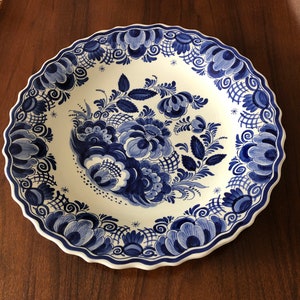 Op de afbeelding: Een grote, witte keramische plaat met een blauw en wit bloemenmotief. De plaat heeft een gegolfde rand en is versierd met ingewikkelde bloemenpatronen.