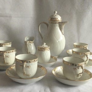 Könnte beinhalten: Ein cremefarbenes Porzellan-Teeservice mit goldenen Verzierungen. Das Set umfasst eine Kaffeekanne, einen Milchgießer, eine Zuckerdose, Untertassen und Tassen. Die Tassen und Untertassen sind kreisförmig angeordnet, mit der Kaffeekanne im Mittelpunkt.