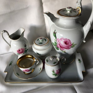 May include: A white porcelain tea set with a rose pattern. The set includes a teapot, creamer, sugar bowl, cup and saucer, and a tray. The set is decorated with pink roses and green leaves.