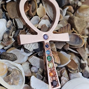 Op de afbeelding: Een ankh-hanger van roségoud met een regenboog van edelstenen die in de verticale balk zijn gezet. De hanger is omringd door schelpen.