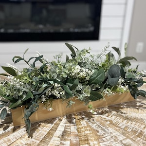 May include: A long, rectangular gold planter filled with a lush arrangement of green and white faux greenery. The planter is sitting on a light wood surface.