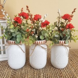 May include: Three white mason jars with red and white striped ribbon and twine, filled with red flowers and green foliage.
