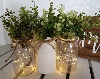 May include: Three mason jars with greenery and fairy lights. Two clear jars and one white jar are tied with twine. The jars are filled with green foliage and small white lights. A wooden sign with white lettering is partially visible.