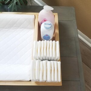May include: A wooden changing table tray with a white quilted changing pad, a stack of diapers, and two bottles of baby lotion. One bottle is pink and says "Johnson's" and the other is blue and says "Johnson's Baby".