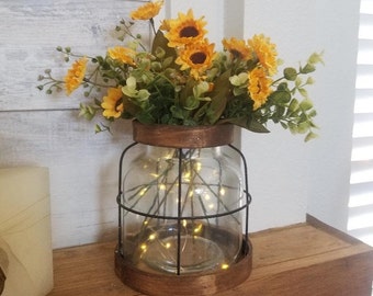 May include: A glass jar with a wire cage and a wooden base, filled with a bouquet of yellow sunflowers and green foliage. The jar is illuminated with string lights.