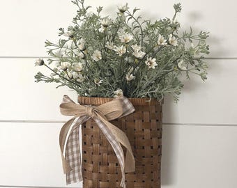 May include: A brown woven basket, filled with white flowers and green foliage, is tied with a tan and white checked ribbon. The rectangular basket hangs on a white, horizontally-slatted wall, creating a rustic, farmhouse aesthetic.