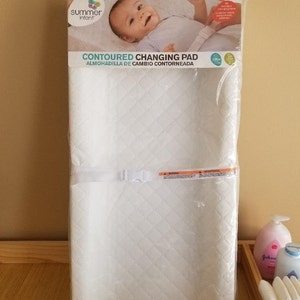 May include: White contoured changing pad with a quilted design and a strap with a buckle. The pad is packaged in plastic and has the words "Contoured Changing Pad" and "Almohadilla de Cambio Contorneada" printed on the front. The pad is made by Summer Infant.