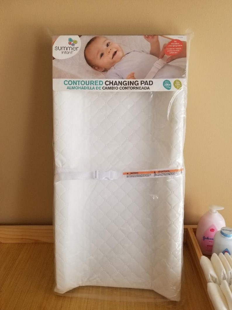 May include: White contoured changing pad with a quilted design and a strap with a buckle. The pad is packaged in plastic and has the words "Contoured Changing Pad" and "Almohadilla de Cambio Contorneada" printed on the front. The pad is made by Summer Infant.