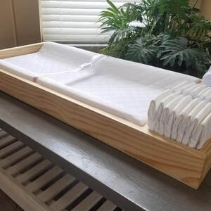 May include: A wooden changing table with a white changing pad, a stack of diapers, and bottles of baby lotion and powder.