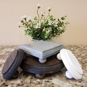 May include: Three wooden tiered display stands with a white, gray, and dark brown finish. Each stand has a round or square top and four small wooden legs. The stands are stacked on top of each other, with the white stand on top, the gray stand in the middle, and the dark brown stand on the bottom.