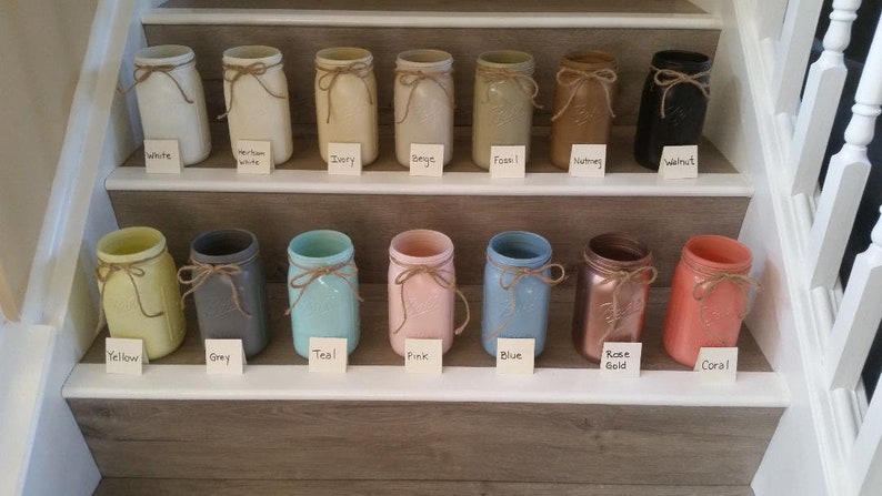 May include: A set of 14 mason jars painted in various colors, including white, ivory, beige, fossil, nutmeg, walnut, yellow, gray, teal, pink, blue, rose gold, and coral. Each jar has a brown twine bow around the lid.