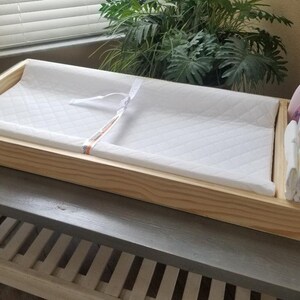 May include: A wooden changing table with a white changing pad and a side compartment for diapers and baby supplies.