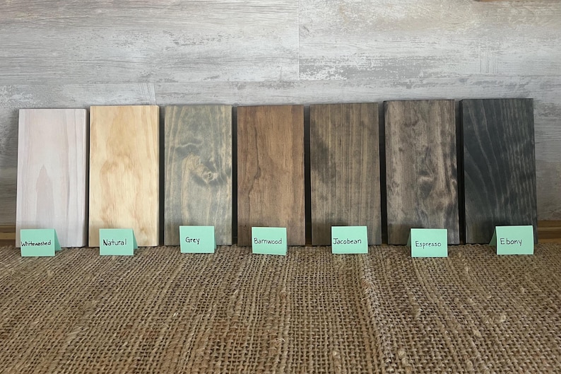 May include: Eight wood samples stained in different colors, including whitewashed, natural, gray, barnwood, jacobean, espresso, and ebony. The samples are arranged horizontally on a burlap surface.