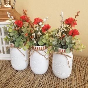 May include: Three white mason jars with red and white striped ribbon and twine, filled with red and white flowers and greenery.