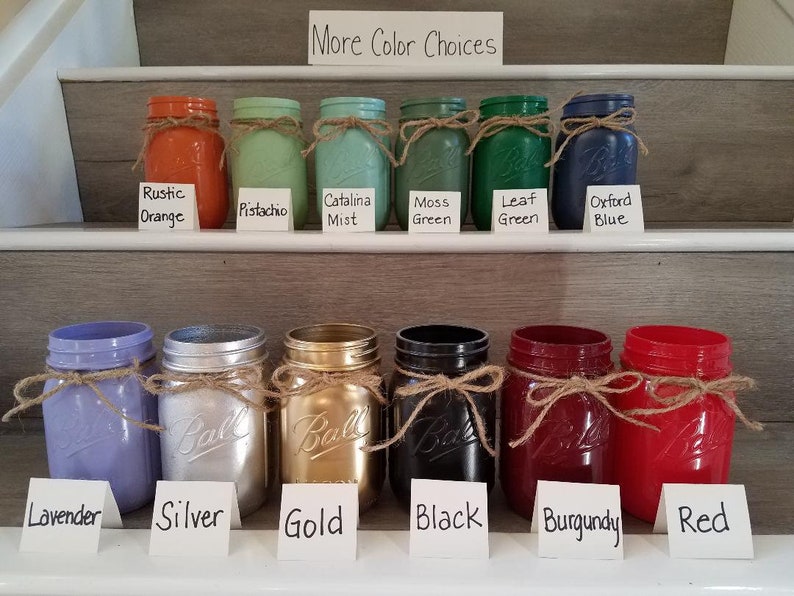 May include: A set of 10 mason jars painted in various colors, including rustic orange, pistachio, Catalina mist, moss green, leaf green, Oxford blue, lavender, silver, gold, black, burgundy, and red. Each jar has a twine bow around the lid. The jars are arranged in two rows on a set of stairs.