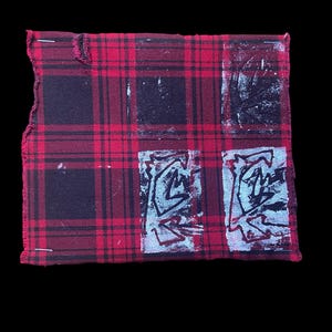 Könnte beinhalten: Ein quadratischer Patch aus rot-schwarz kariertem Stoff mit vier dunklen Rechtecken und zwei hellblau-schwarzen abstrakten Designs. Die Kanten des Stoffes sind ausgefranst und angesteckt.